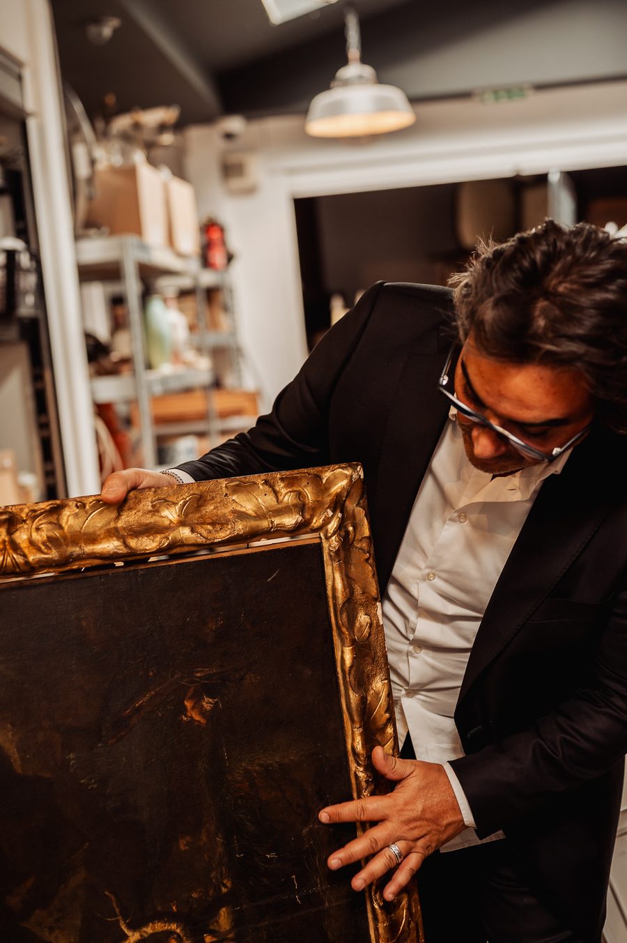 Éric Benhamou examine une huile sur toile ancienne encrassée, la main posée sur son cadre en bois doré sculpté de feuillages.
