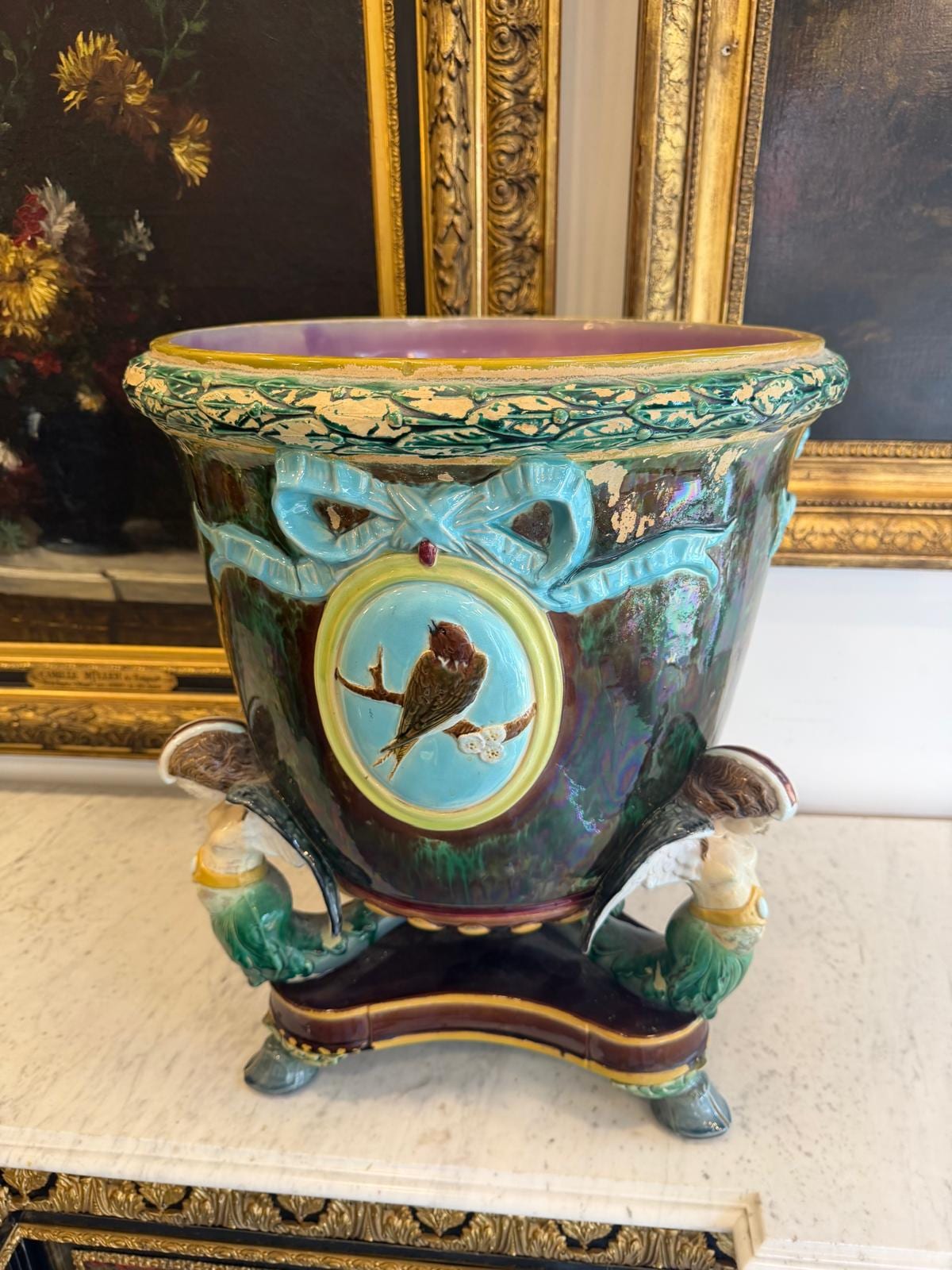 Jardinière en barbotine à fond vert et brun flammé, médaillon turquoise à l'oiseau, posée sur trois figures ailées et un socle tripode, sur une console en marbre devant des tableaux encadrés d'or