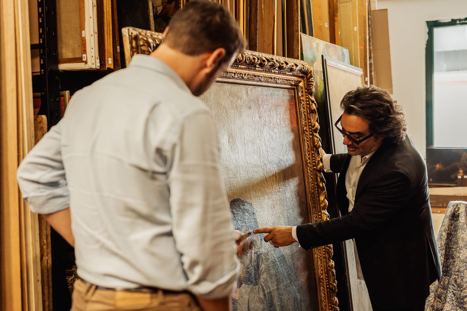 Éric BENHAMOU montre à un visiteur un détail de la toile, un grand portrait à l'huile dans un cadre doré à feuillages, dans la réserve des tableaux
