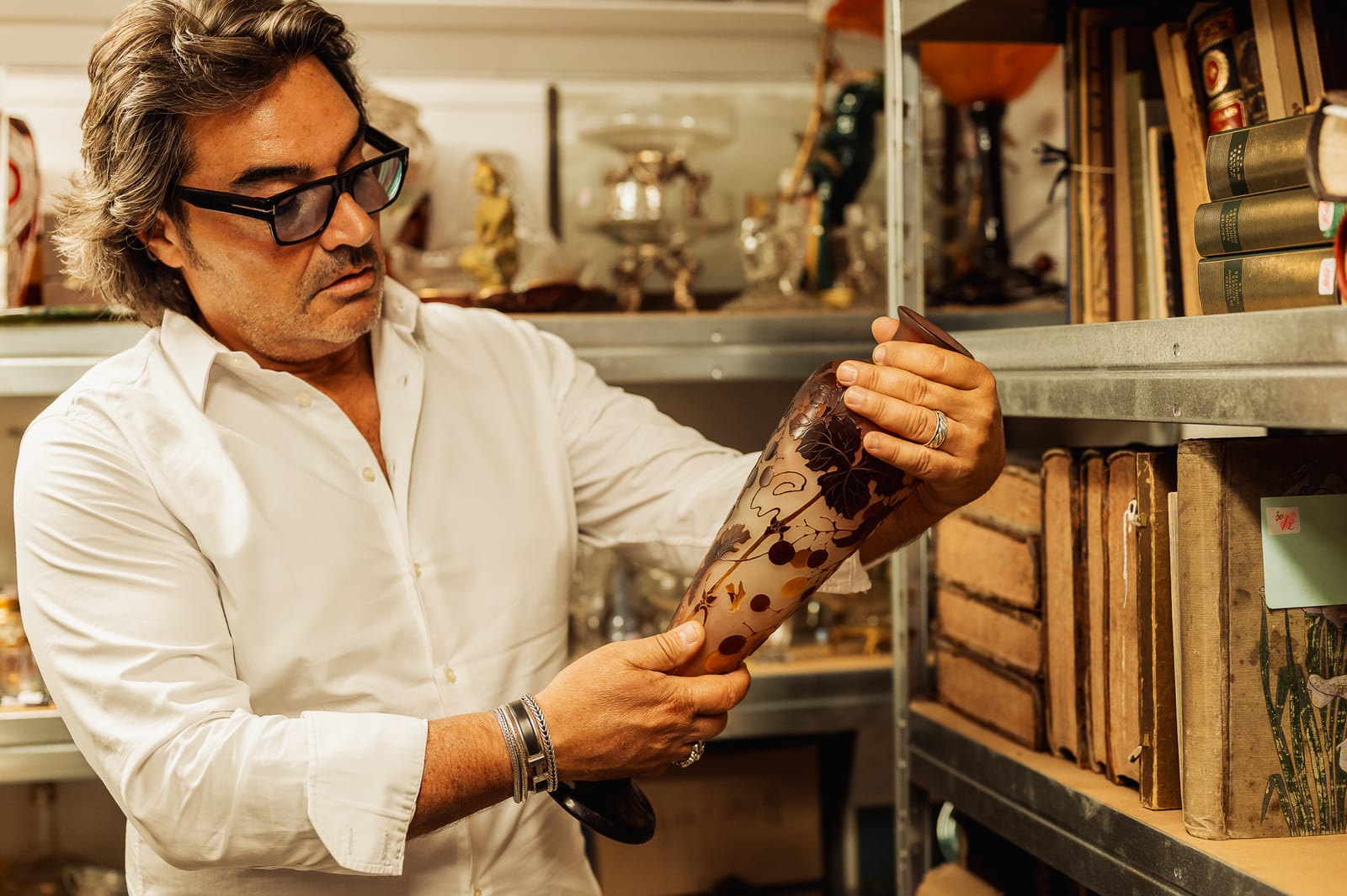 Éric BENHAMOU, antiquaire, examine un grand vase cornet en verre multicouche à décor de baies brunes, entre des étagères chargées d'objets